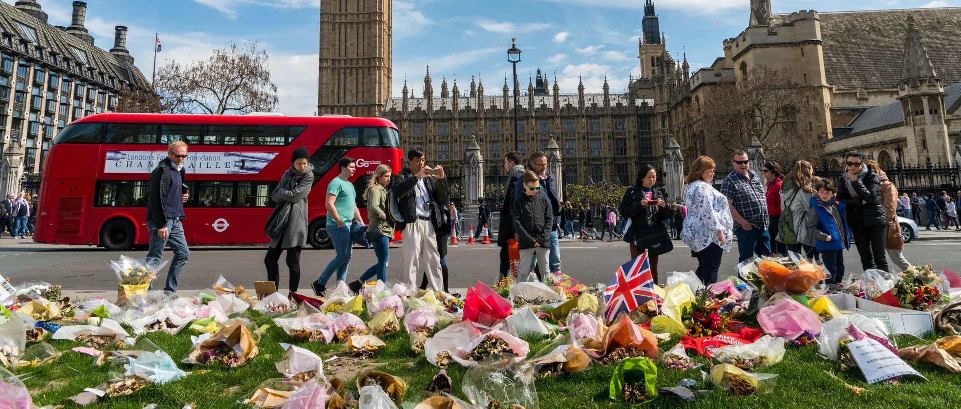 Flowers and a Union flag laid on the grass opposite the Houses of Parliament, with people walking past and a red bus behind.
