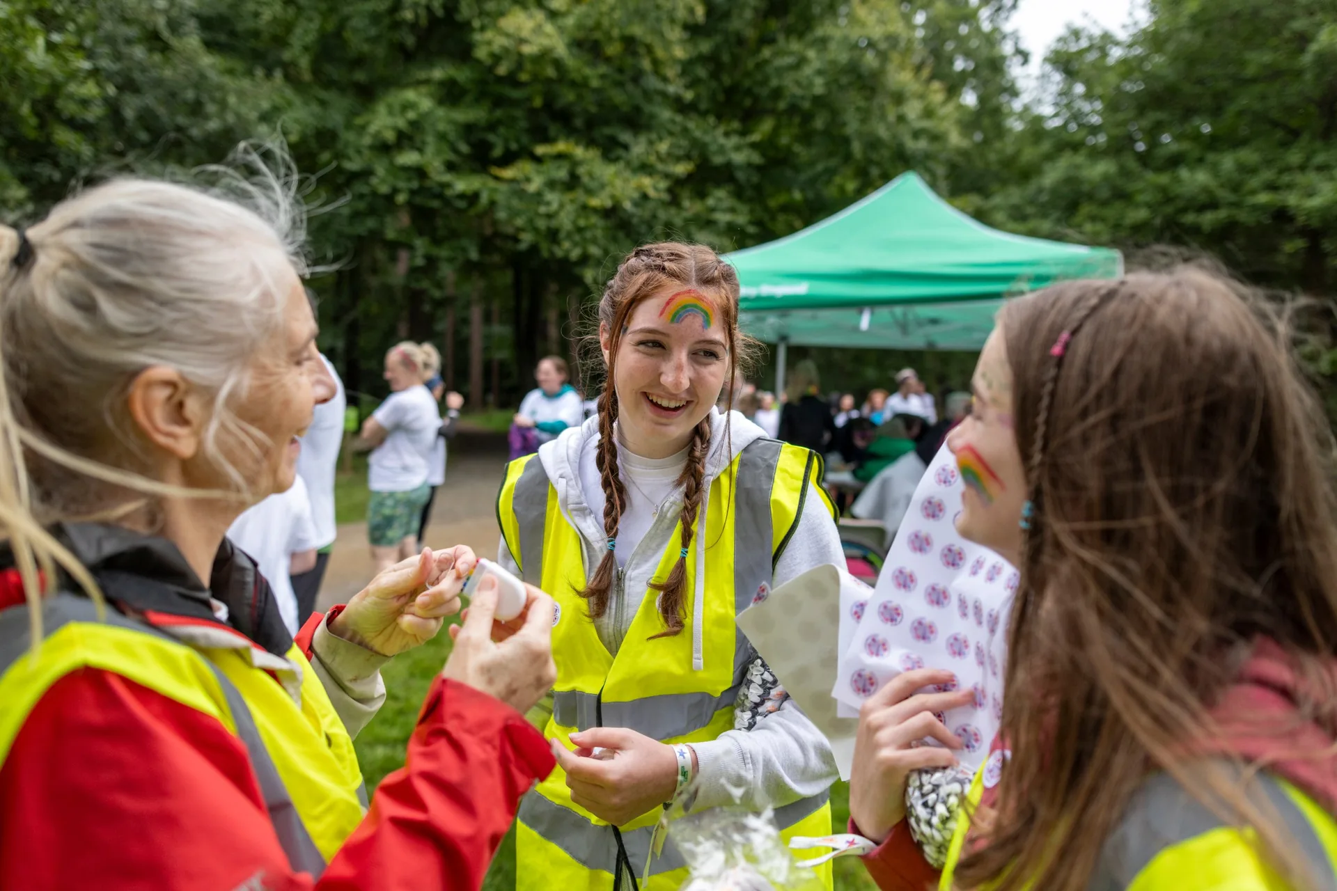 Three people in high-visibility vests laugh together at an outdoor event in a wood. The woman in the middle has a rainbow painted on her forehead.