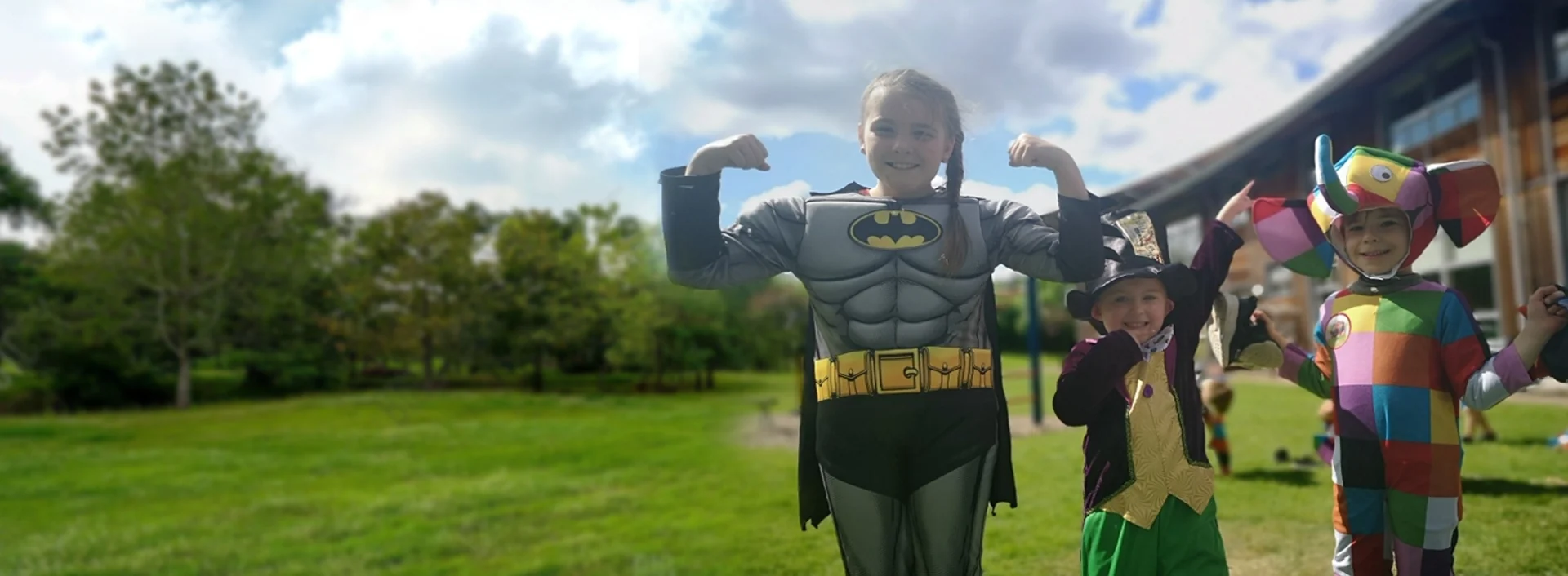 Three smiling children in fancy dress on a sunny field. The girl at the front flexes her arms in a Batman costume.