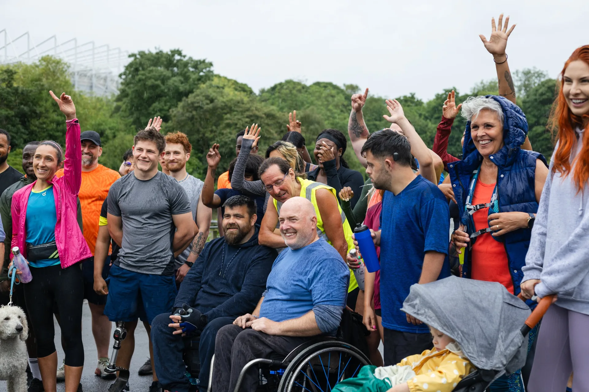 A smiling group of people in sportswear, some waving, gathered outdoors on a rainy day. One man sits in a wheelchair at the front.