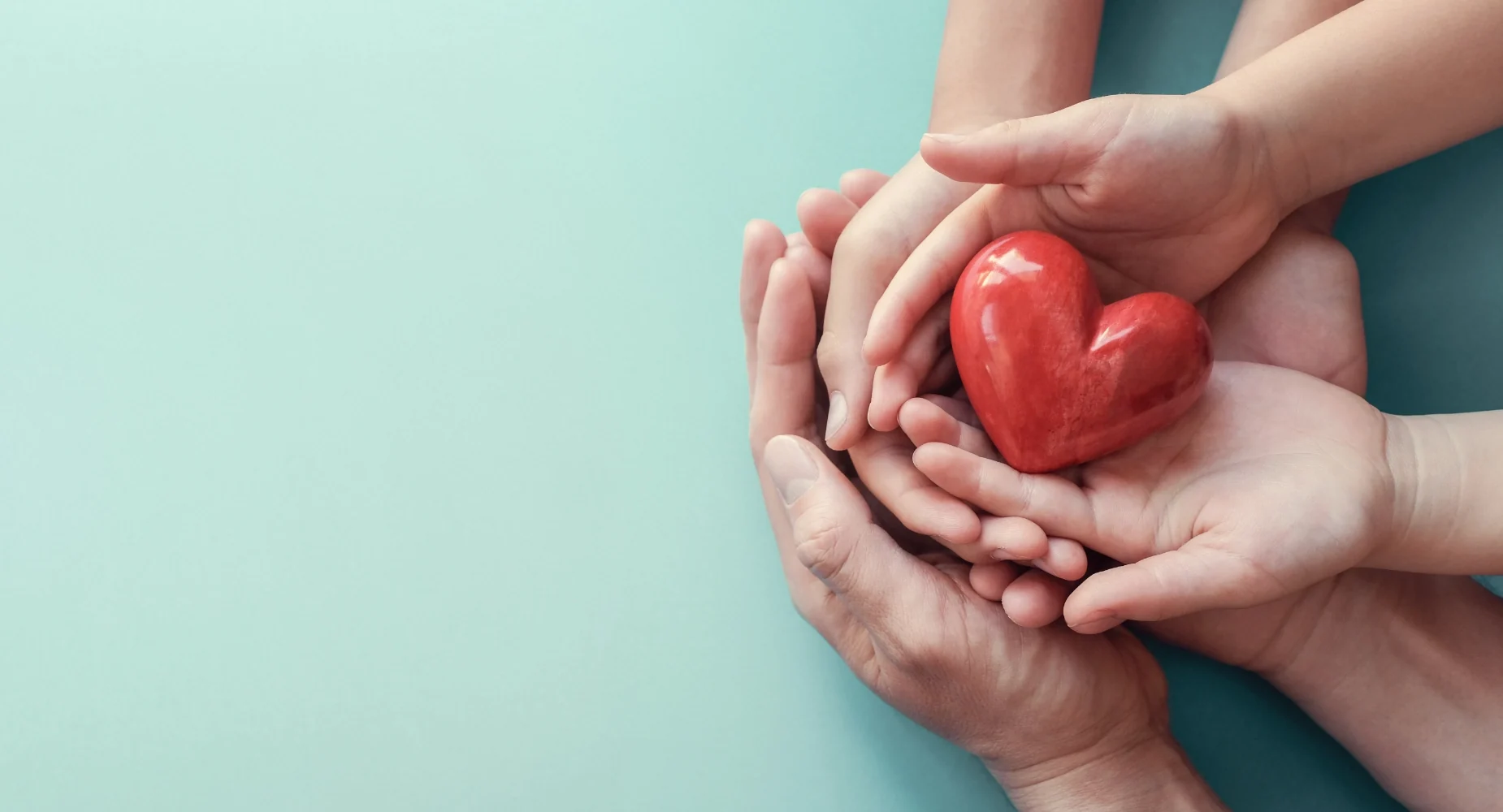 An adult's and a child's hands cupped together, holding a red heart, on a pale blue-green background.
