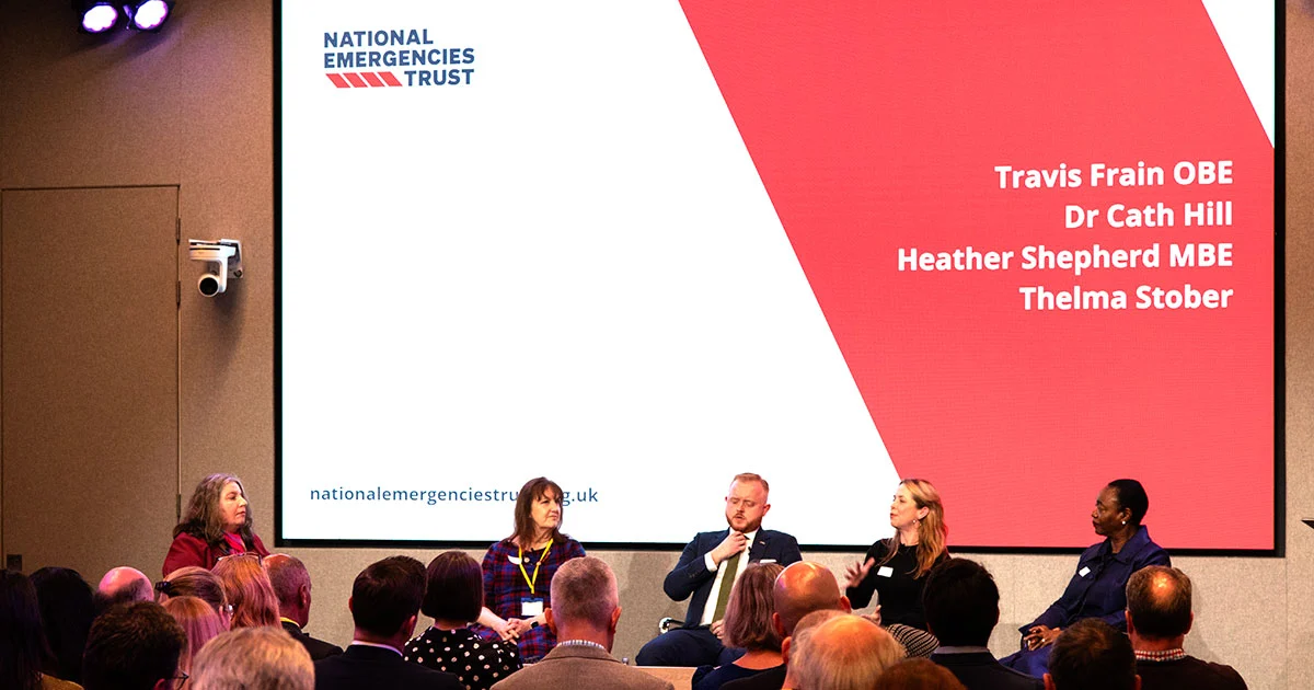 Five panellists on stage at our event, in front of an audience. The screen names Travis Frain OBE, Dr Cath Hill, Heather Shepherd MBE and Thelma Stober.