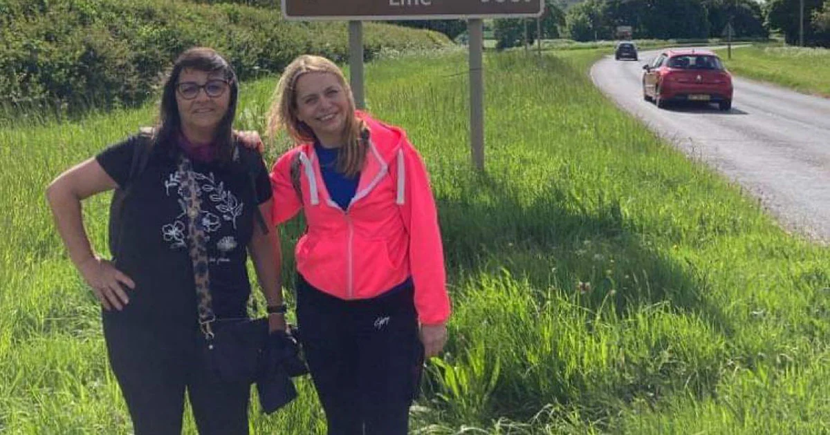 Two women smiling on a grass verge beside a country road.